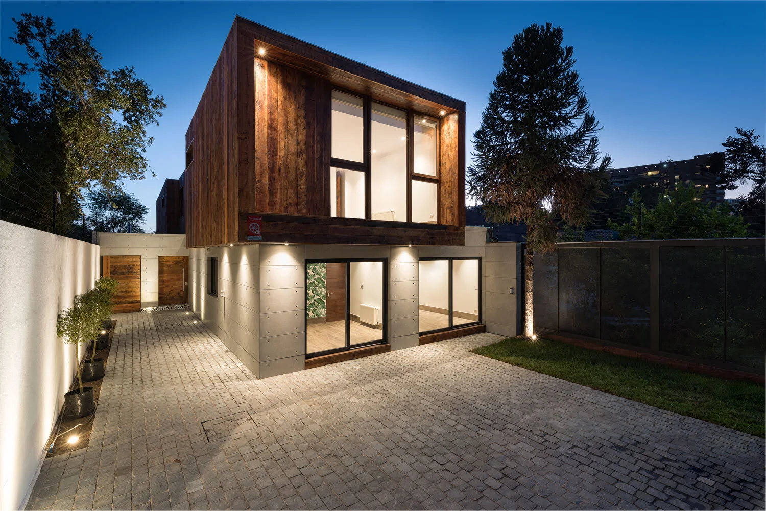 Casa de dos pisos con volumen superior revestido en madera y primer piso en placas de hormigón, iluminada al anochecer