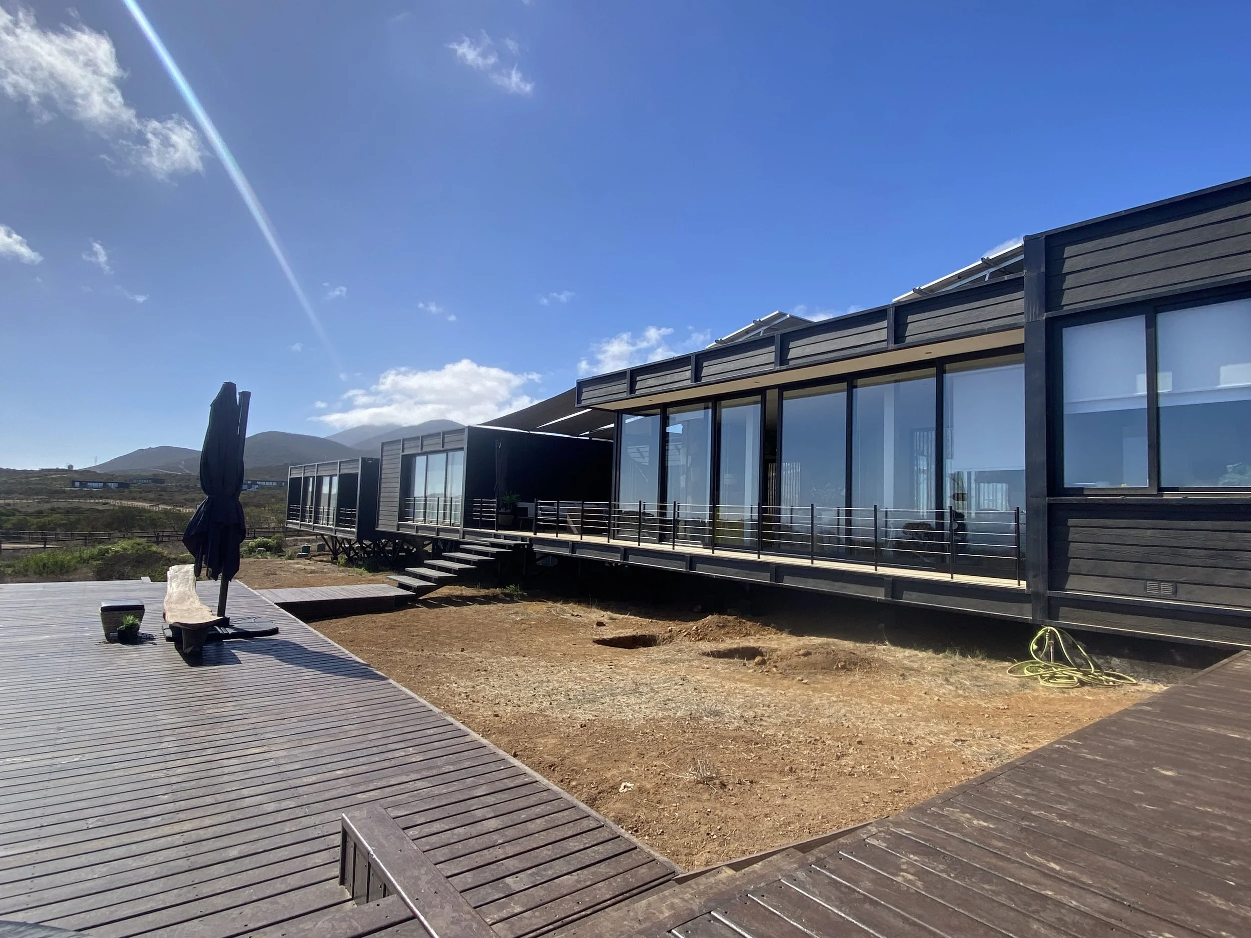 Casa CD en Los Molles: volúmenes oscuros con grandes ventanales y terraza de madera frente a los cerros