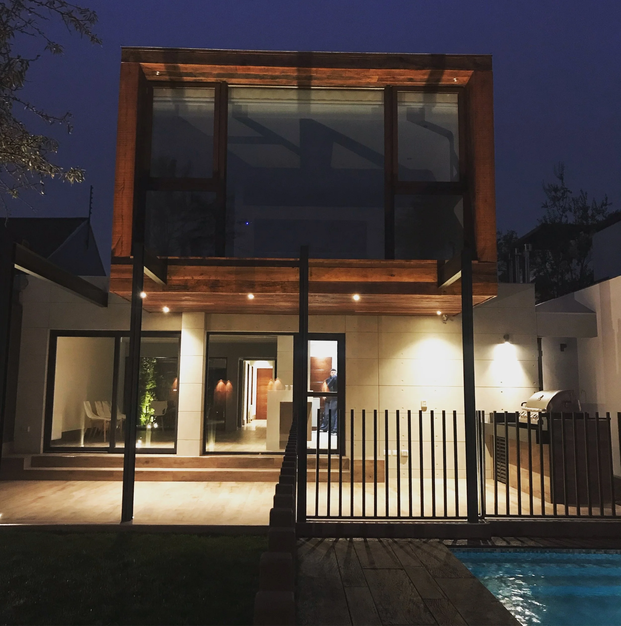 Fachada posterior iluminada de noche, con volumen de madera en voladizo, terraza y piscina