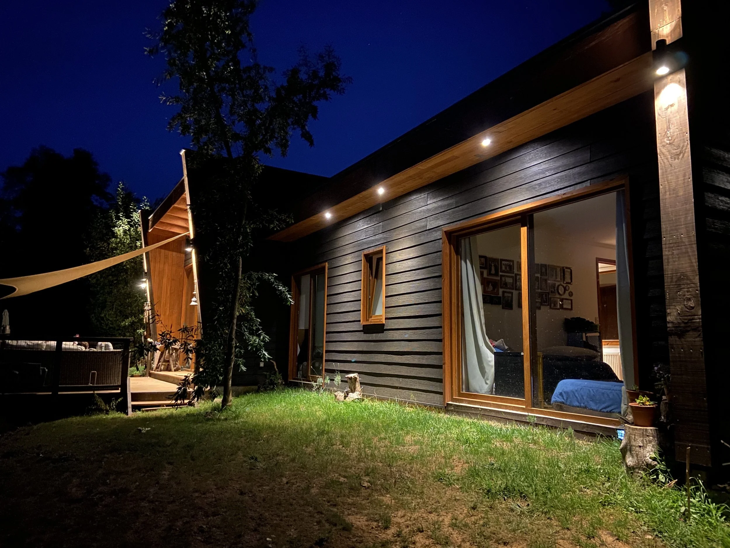 Casa con revestimiento oscuro y marcos de madera, iluminada de noche junto a un jardín